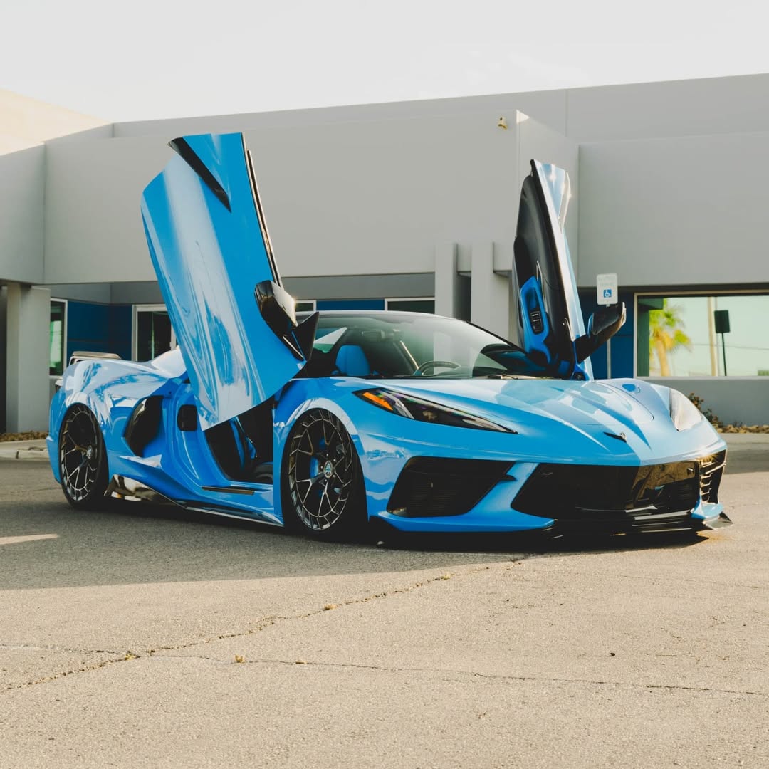 Blue Corvette C8 with custom aftermarket wheels and butterfly doors in a modern outdoor setting.